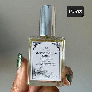 May include: A rectangular perfume bottle with a silver cap. The label reads "Marshmallow Musk" and "Extrait de Parfum". The bottle is 0.5 oz / 15 ml. The bottle is held by a person with green nail polish.