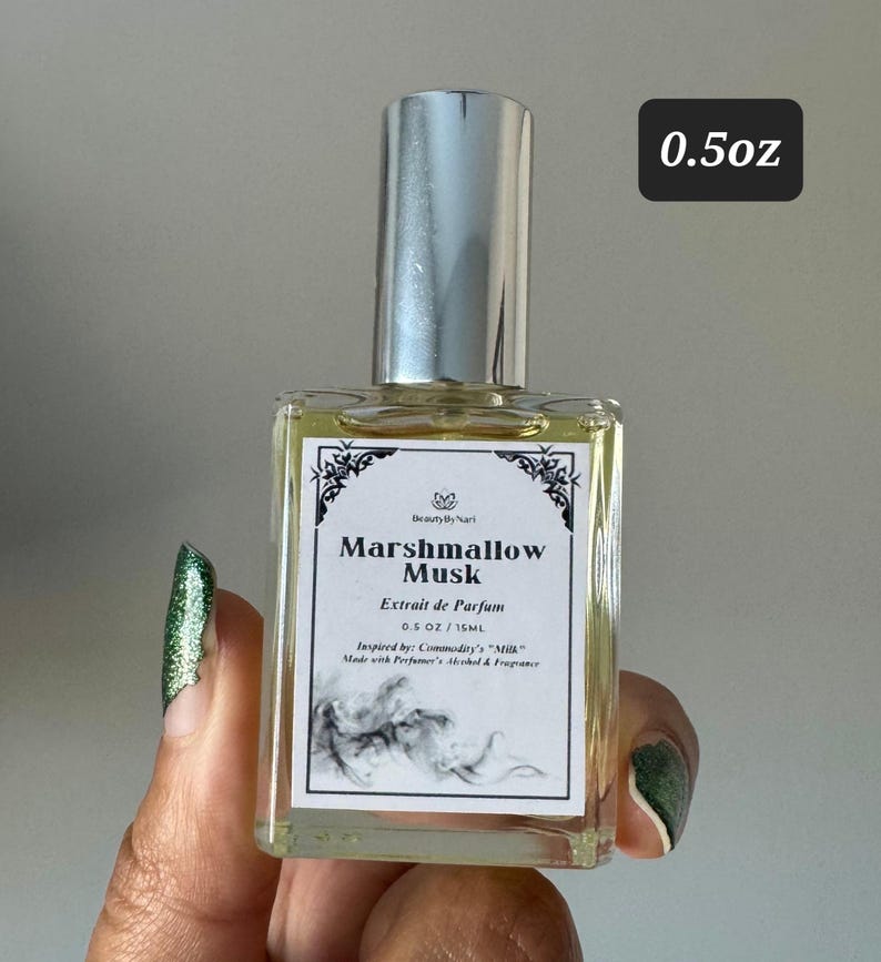May include: A rectangular perfume bottle with a silver cap. The label reads "Marshmallow Musk" and "Extrait de Parfum". The bottle is 0.5 oz / 15 ml. The bottle is held by a person with green nail polish.