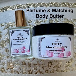 May include: A perfume bottle and a jar of body butter with the text "Puffy Marshmallow". The perfume bottle has a gold cap and a label with the text "Extrait de Parfum". The body butter jar has a black lid and a label with the text "Whipped Body Buttercream".