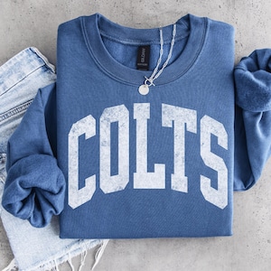 Puede incluir: Una sudadera azul de cuello redondo con la palabra COLTS en letras grandes de bloque blanco desgastado en el pecho. La prenda se combina con unos vaqueros azul claro y un collar de cadena plateada con un pequeño colgante circular.