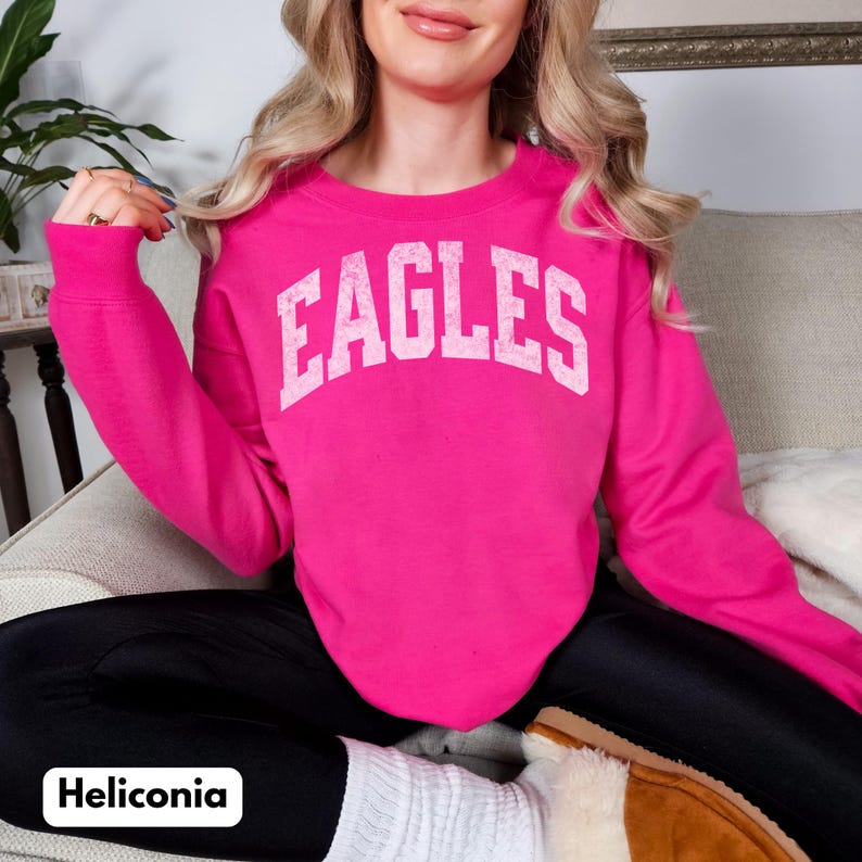 May include: A vibrant pink crew neck sweatshirt featuring the word "EAGLES" in white capital letters. The image also shows black leggings and white socks. The sweatshirt is a casual style.