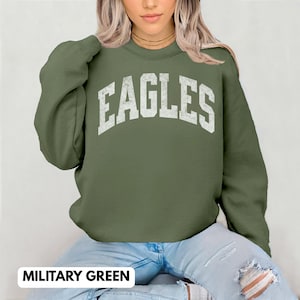 May include: A military green crew neck sweatshirt featuring the word "EAGLES" in distressed white lettering. The sweatshirt is made of a soft material. The text "MILITARY GREEN" is displayed at the bottom of the image.