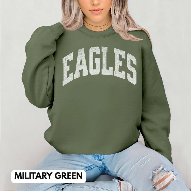 May include: A military green crew neck sweatshirt featuring the word "EAGLES" in distressed white lettering. The sweatshirt is made of a soft material. The text "MILITARY GREEN" is displayed at the bottom of the image.