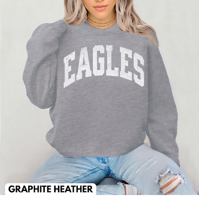 May include: A grey sweatshirt with the word "EAGLES" in large, distressed white letters across the chest. The sweatshirt has long sleeves and a crew neck. The text "GRAPHITE HEATHER" is at the bottom of the image.