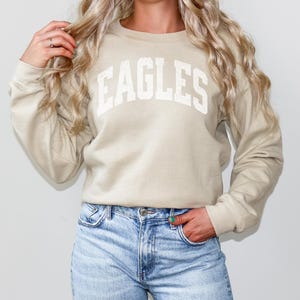 May include: A sand-coloured sweatshirt with the word "EAGLES" in white block letters. The long-sleeved crew neck top is paired with light blue denim jeans. The word "Sand" is printed in a white box at the bottom.