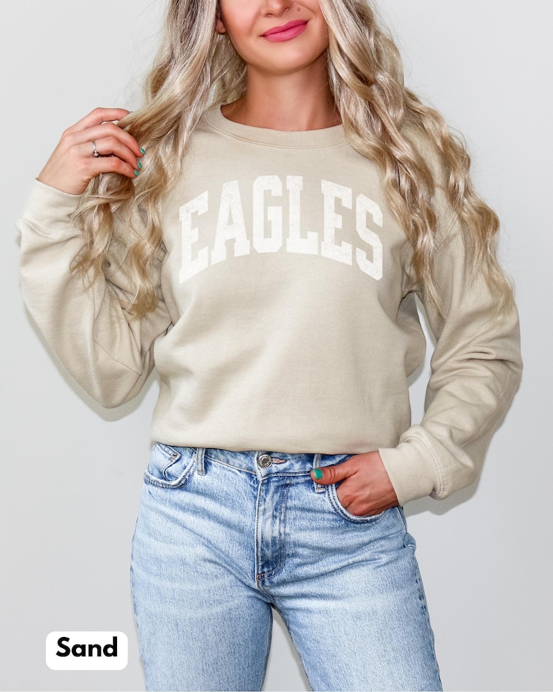 May include: A sand-coloured sweatshirt with the word "EAGLES" in white block letters. The long-sleeved crew neck top is paired with light blue denim jeans. The word "Sand" is printed in a white box at the bottom.