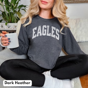 May include: Dark grey sweatshirt featuring the word "EAGLES" in white, arched lettering. The sweatshirt is paired with black leggings and white socks. A white mug is held in the hand.