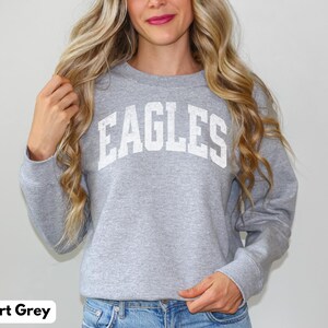 May include: Grey sweatshirt with the word "EAGLES" in white block letters. The long-sleeved crew neck sweatshirt is made of a soft material. The model is wearing blue jeans.