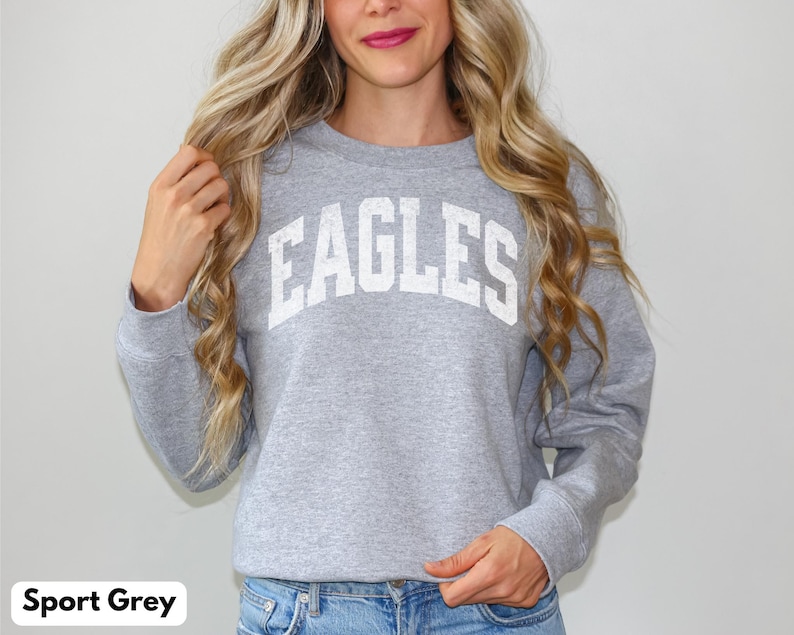 May include: Grey sweatshirt with the word "EAGLES" in white block letters. The long-sleeved crew neck sweatshirt is made of a soft material. The model is wearing blue jeans.