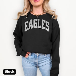 May include: Black crew neck sweatshirt featuring the word "EAGLES" in distressed white lettering. The long-sleeved sweatshirt is worn with blue jeans. The word "Black" is in a white box in the lower left corner of the image.