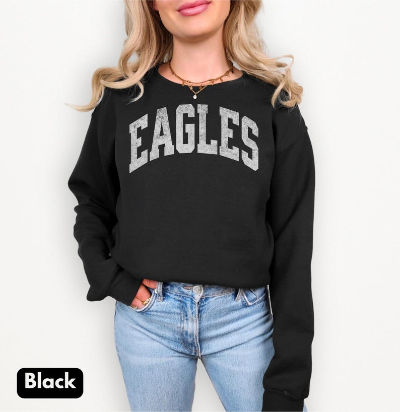 May include: Black crew neck sweatshirt featuring the word "EAGLES" in distressed white lettering. The long-sleeved sweatshirt is worn with blue jeans. The word "Black" is in a white box in the lower left corner of the image.