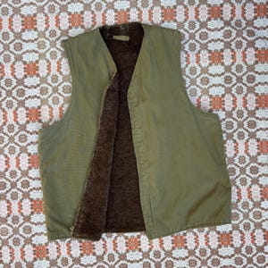 Op de afbeelding: Olijfgroen vest met een bruine, donzige binnenkant. Het vest heeft een volledige ritssluiting en een V-hals. De buitenkant lijkt van een duurzame stof te zijn, en het vest is waarschijnlijk ontworpen voor warmte. Het vest ligt plat.