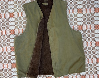 Può includere: Gilet verde oliva con interno marrone e soffice. Il gilet ha una chiusura con cerniera completa e un design a collo a V. Il materiale esterno sembra essere un tessuto resistente e il gilet è probabilmente progettato per il calore. Il gilet è piatto.