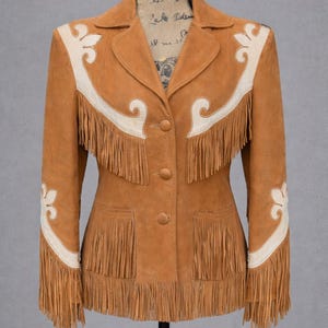 Puede incluir: Chaqueta de ante marrón claro con detalles decorativos color crema y flecos. La chaqueta presenta un cuello con solapas, dos bolsillos delanteros y cierres de botón. Los flecos están en el dobladillo, los bolsillos y las mangas.