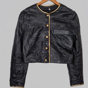 May include: Black leather jacket with gold trim and buttons. The jacket has a round collar and a small pocket. The jacket is hanging on a wooden hanger.
