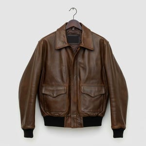 Könnte beinhalten: Braune Leder-Bomberjacke mit klassischem Design. Die Jacke hat einen Kragen, zwei Vordertaschen und schwarze gerippte Bündchen und Saum. Sie hängt an einem Holzbügel.