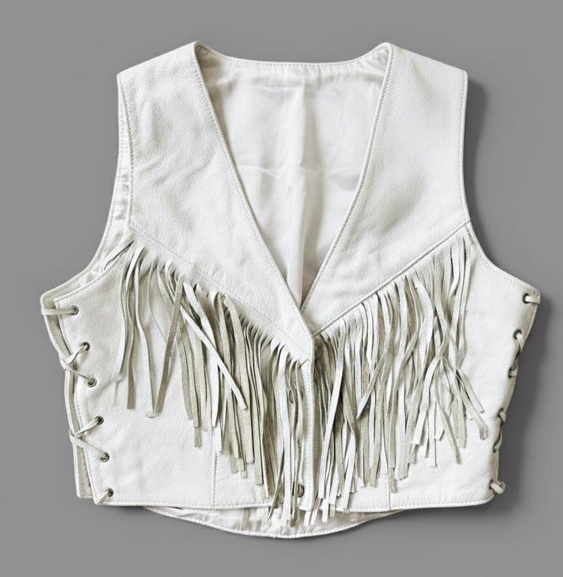 May include: A white leather vest with a V-neck and fringe detailing. The vest has a lace-up design on the sides and a smooth, solid back. The fringe is long and hangs from the front of the vest.