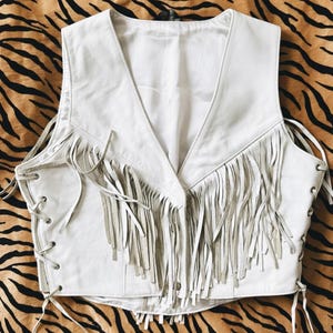 May include: A white leather vest with a V-neck and fringe detailing. The vest has a lace-up design on the sides and a button closure. The fringe is a lighter shade of white. The vest is a stylish piece of apparel.