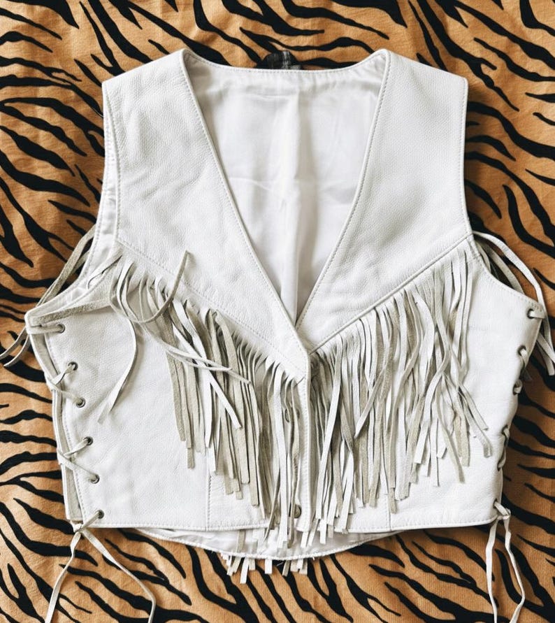 May include: A white leather vest with a V-neck and fringe detailing. The vest has a lace-up design on the sides and a button closure. The fringe is a lighter shade of white. The vest is a stylish piece of apparel.