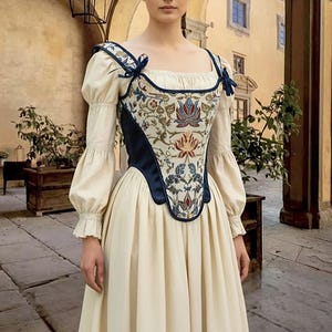Op de afbeelding: Cr&egrave;mekleurige historische jurk met een bloemen geborduurd lijfje. Het lijfje heeft marineblauwe randen en accenten. De mouwen zijn lang en poffend, en de rok is wijd. De jurk doet denken aan de mode uit de Renaissance.