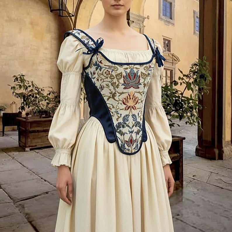 Op de afbeelding: Cr&egrave;mekleurige historische jurk met een bloemen geborduurd lijfje. Het lijfje heeft marineblauwe randen en accenten. De mouwen zijn lang en poffend, en de rok is wijd. De jurk doet denken aan de mode uit de Renaissance.