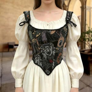 May include: A black and gray tapestry corset with a witch-themed design, featuring a pointed neckline and black straps with bows. The corset is worn over a cream-colored blouse with puffy sleeves and a white dress.