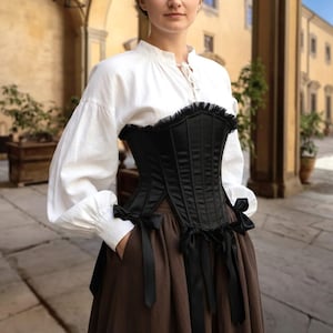 Op de afbeelding: Een zwart korset met een gefranjerde top, gecombineerd met een witte blouse en een bruine rok. Het korset heeft verticale naden en zwarte strikbanden. De blouse heeft lange, pofmouwen.