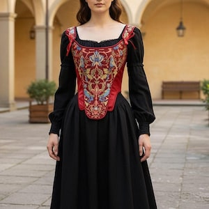 Op de afbeelding: Zwarte jurk met lange mouwen en een vierkante halslijn. Het lijfje is rood met een geborduurd bloemmotief in goud, blauw en groen. Rode linten accentueren de schouders. De jurk heeft een volle rok, in historische stijl.