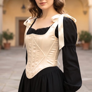May include: A cream-colored corset with vertical boning and ribbon shoulder ties, worn over a black long-sleeved top with a square neckline and gathered sleeves. The corset features a structured bodice with a pointed hemline and satin finish.