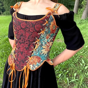 Op de afbeelding: Een korsettop met een gespleten ontwerp, voorzien van een rood rozenpatroon aan de ene kant en een veelkleurige bloemenprint aan de andere kant. Het kledingstuk is afgewerkt met oranje lintveters door zilveren oogjes, oranje biezen en schouderstrikken.