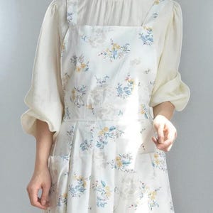 May include: White apron with a floral pattern featuring yellow and blue flowers. The apron has a square neckline, two front pockets, and wide shoulder straps. The apron is worn over a white blouse with puffy sleeves.