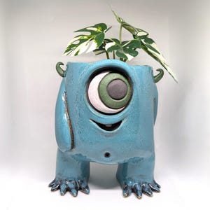 Op de afbeelding: Een blauwe keramische plantenpot in de vorm van een eenogig monster met twee benen, klauwvoeten en kleine hoorns. Het monster heeft een groot centraal oog met een groene iris en een kleine mond. Een groen-witte plant groeit uit de opening bovenop.