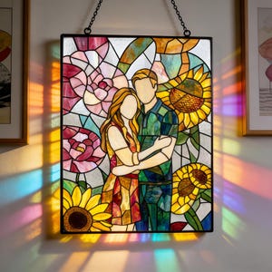 May include: A rectangular stained glass panel featuring a faceless couple embracing, surrounded by vibrant pink flowers and yellow sunflowers. The piece is framed in black metal and hangs from a chain, casting colorful light patterns on the wall.