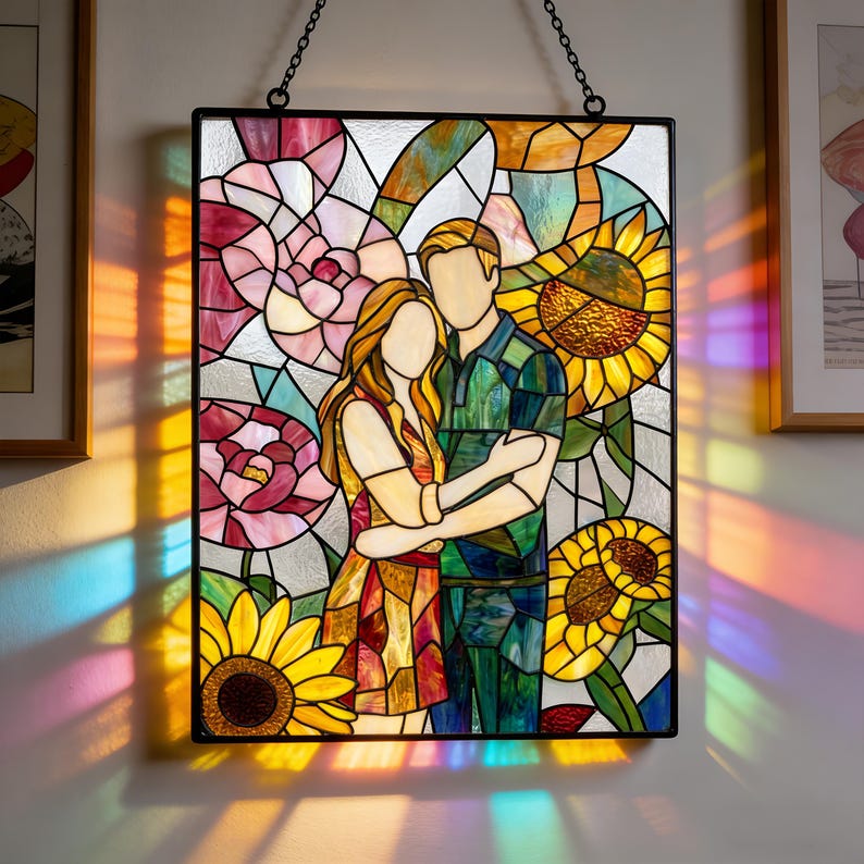 May include: A rectangular stained glass panel featuring a faceless couple embracing, surrounded by vibrant pink flowers and yellow sunflowers. The piece is framed in black metal and hangs from a chain, casting colorful light patterns on the wall.