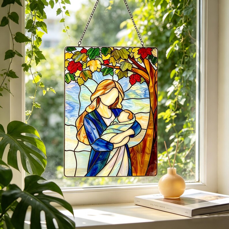 May include: A rectangular stained glass suncatcher featuring a mother figure with long blonde hair holding a swaddled infant. The scene is set under a tree with red, orange, and green leaves against a blue sky, suspended by a metal chain.