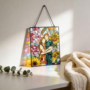 May include: A rectangular stained glass panel featuring a couple embracing, surrounded by vibrant sunflowers and pink flowers. The artwork is framed in black metal and suspended by a black chain, casting colorful light reflections on the surface.