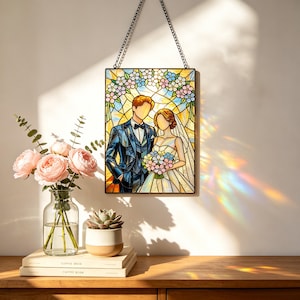 May include: A rectangular stained glass suncatcher featuring a wedding couple in a suit and gown, surrounded by floral patterns in blue, pink, and yellow. The piece is framed in black metal and hangs from a black chain, casting colorful light refractions.