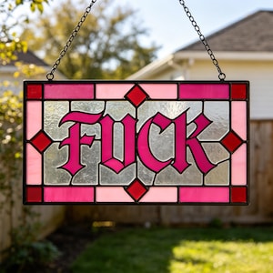 May include: A rectangular stained glass panel featuring the word FUCK in bold, dark pink gothic lettering. The piece is framed by a grid of clear, light pink, and deep red glass segments, held together by black metal cames and suspended by a chain.