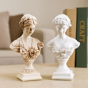 May include: Two classical style busts of women. One is a light beige colour, the other is white. Both have detailed facial features, curly hair, and are wearing draped garments. They are displayed on a wooden table.