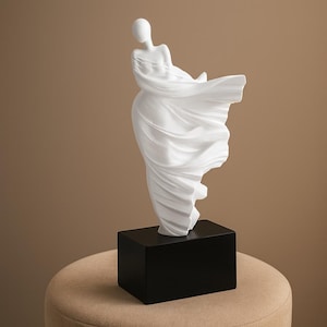 May include: A white sculpture of a draped figure, mounted on a black rectangular base. The figure's form is flowing, with fabric-like details. The sculpture is set against a neutral background.