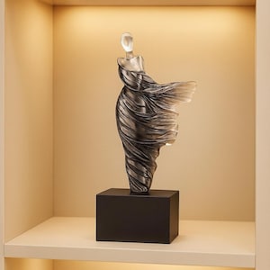 Puede incluir: Una escultura de vidrio transparente de una figura con un vestido fluido, montada sobre una base rectangular negra. La forma de la figura es detallada, con el vestido que parece ondear en el viento. La escultura se exhibe en un estante blanco.