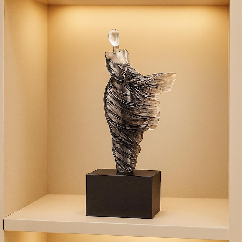 Puede incluir: Una escultura de vidrio transparente de una figura con un vestido fluido, montada sobre una base rectangular negra. La forma de la figura es detallada, con el vestido que parece ondear en el viento. La escultura se exhibe en un estante blanco.