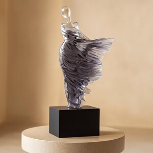 Puede incluir: Una escultura transl&uacute;cida de una figura con un vestido fluido. La figura est&aacute; montada sobre una base rectangular negra, que se asienta sobre un pedestal redondo beige. El vestido parece ser de color gris ahumado, con la luz reflej&aacute;ndose en los pliegues.