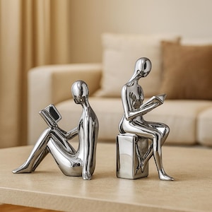 May include: Two silver sculptures of figures reading books. One figure is seated on a cube, the other is reclining. The sculptures have a smooth, reflective finish and are on a light-coloured surface.