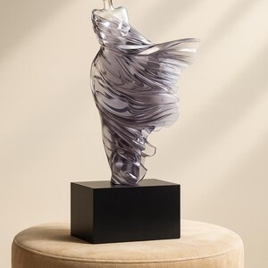 Puede incluir: Una escultura transl&uacute;cida de una figura con un vestido fluido, montada sobre una base rectangular negra. La escultura tiene una calidad suave y et&eacute;rea, con la tela que parece ondear en el viento. La base est&aacute; sobre un taburete tapizado beige.
