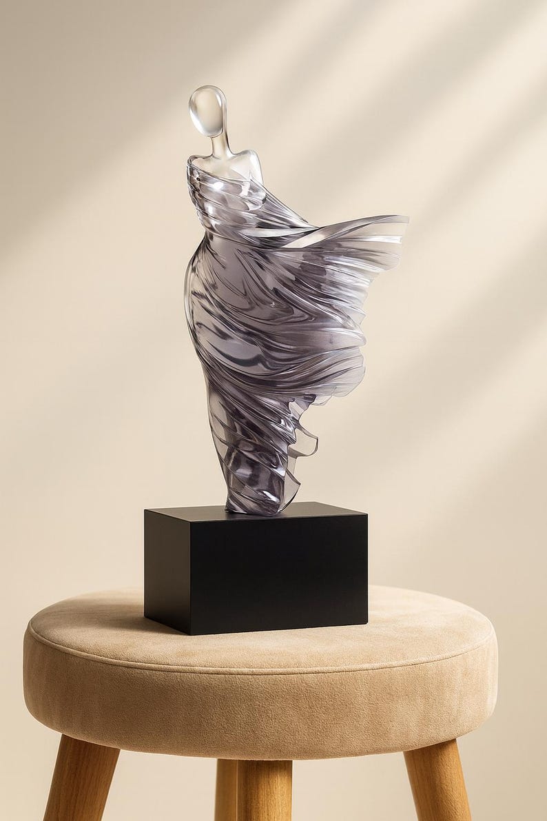 Puede incluir: Una escultura transl&uacute;cida de una figura con un vestido fluido, montada sobre una base rectangular negra. La escultura tiene una calidad suave y et&eacute;rea, con la tela que parece ondear en el viento. La base est&aacute; sobre un taburete tapizado beige.