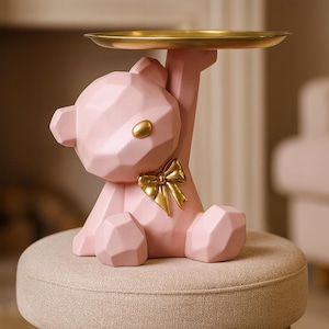 May include: A pink geometric bear-shaped decorative tray with a gold-coloured tray top and bow. The bear has gold-coloured accents on the eyes and bow. The tray is sitting on a round, light-coloured stool.