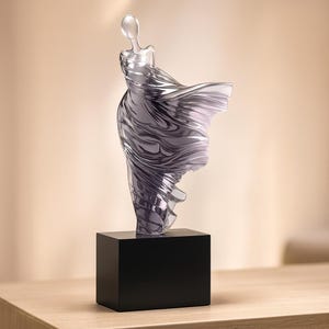 Könnte beinhalten: Eine durchscheinende Skulptur einer Figur in einem fließenden Kleid, montiert auf einem schwarzen rechteckigen Sockel. Das Kleid scheint sich zu bewegen, mit wirbelnden Falten und einem Farbverlauf aus Grau- und Silbertönen. Die Skulptur ist ein dekoratives Kunstwerk.