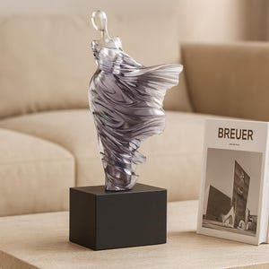 Puede incluir: Una escultura abstracta transparente de una figura con un vestido fluido, montada sobre una base cuadrada negra. La escultura est&aacute; junto a un libro con la palabra "BREUER" en la portada.