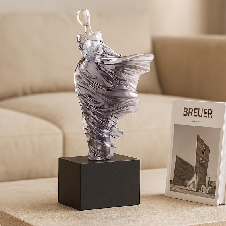 Puede incluir: Una escultura abstracta transparente de una figura con un vestido fluido, montada sobre una base cuadrada negra. La escultura est&aacute; junto a un libro con la palabra "BREUER" en la portada.